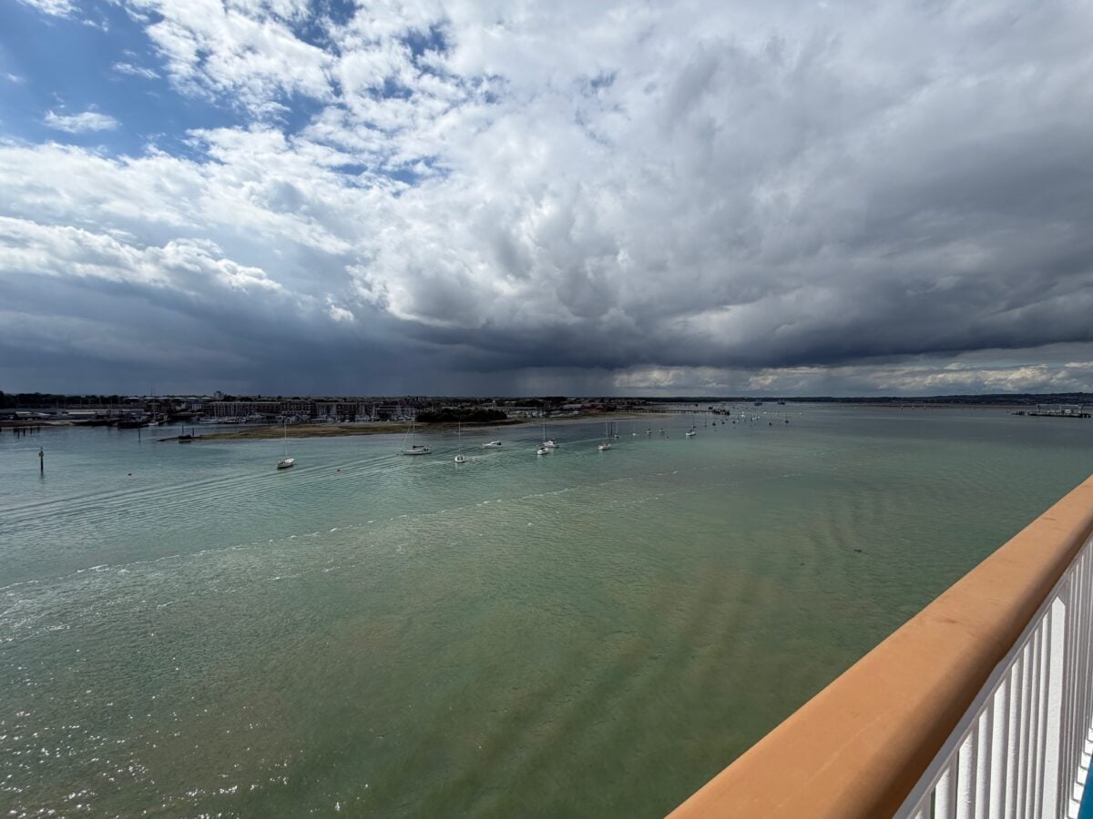 View from a bridge overlooking a calm, greenish body of water with small boats. The sky is partly cloudy, with dark storm clouds gathering on the horizon. A town is visible in the distance.