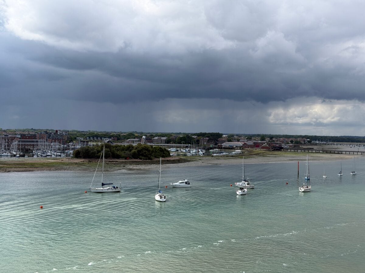 Sailboats anchored on calm water near a shoreline with buildings and trees, under a cloudy sky with dark, looming storm clouds in the distance.