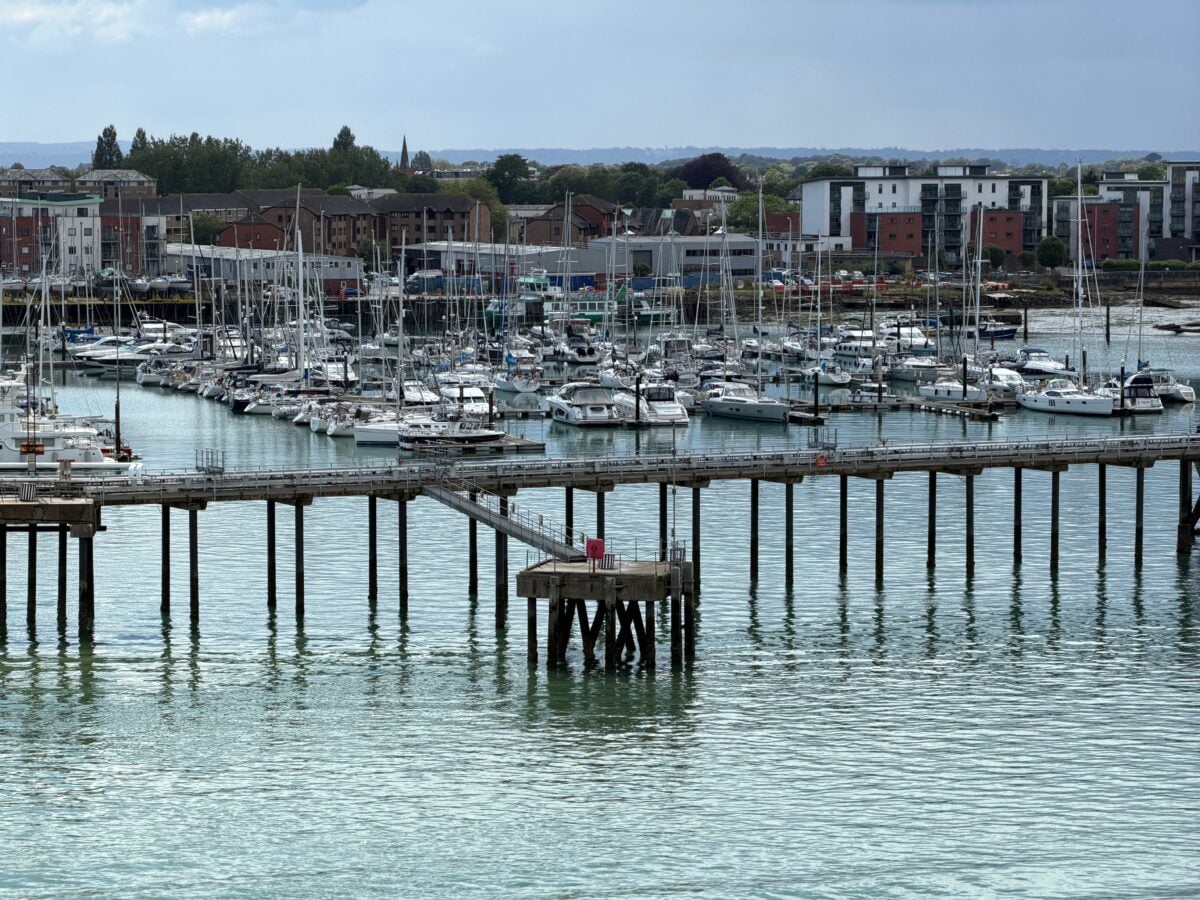 A marina filled with docked sailboats and yachts, with a long wooden pier extending over calm water. In the background are modern buildings and trees under a cloudy sky.