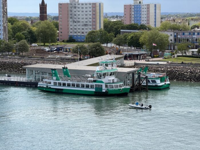 Two green and white ferries are docked at a waterside terminal, with a small motorboat passing in front. High-rise buildings, trees, and a clock tower are visible in the background.