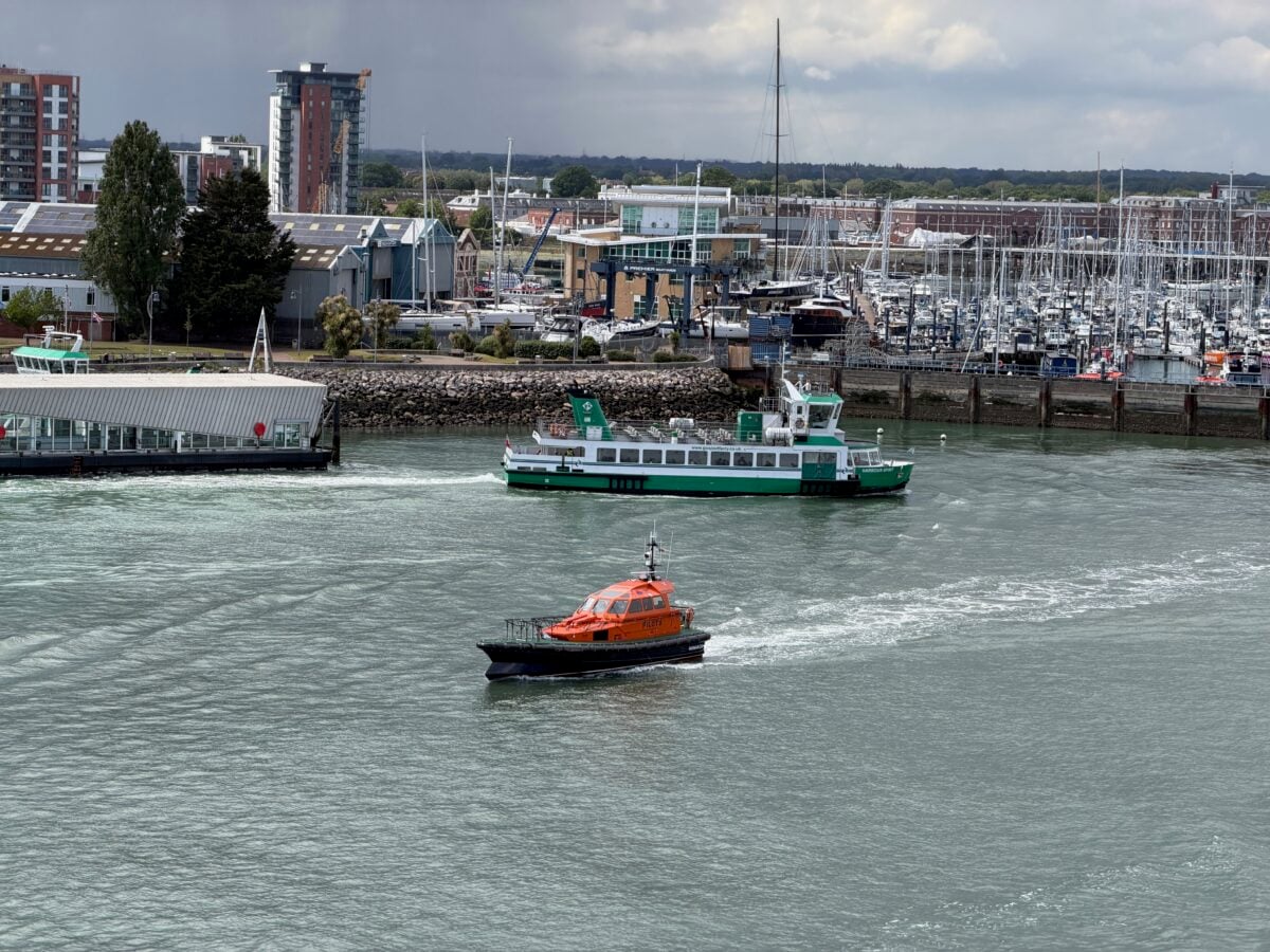 A green and white ferry and an orange and black pilot boat travel on a river near a marina full of sailboats, with buildings and trees visible in the background under a cloudy sky.