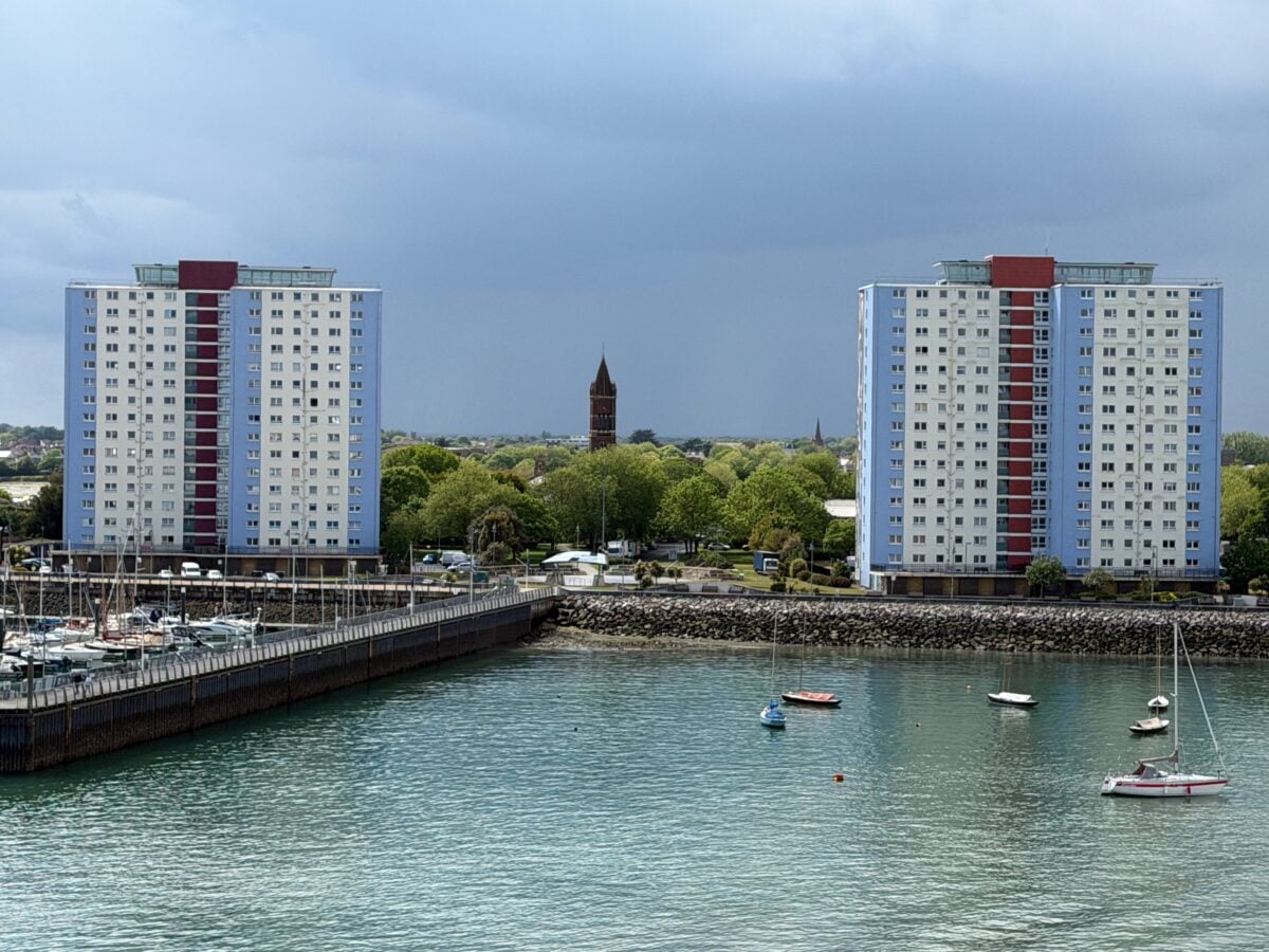 Two tall apartment buildings with red and blue accents stand on either side of a marina with small boats. Trees and a church tower appear in the background under a cloudy sky.