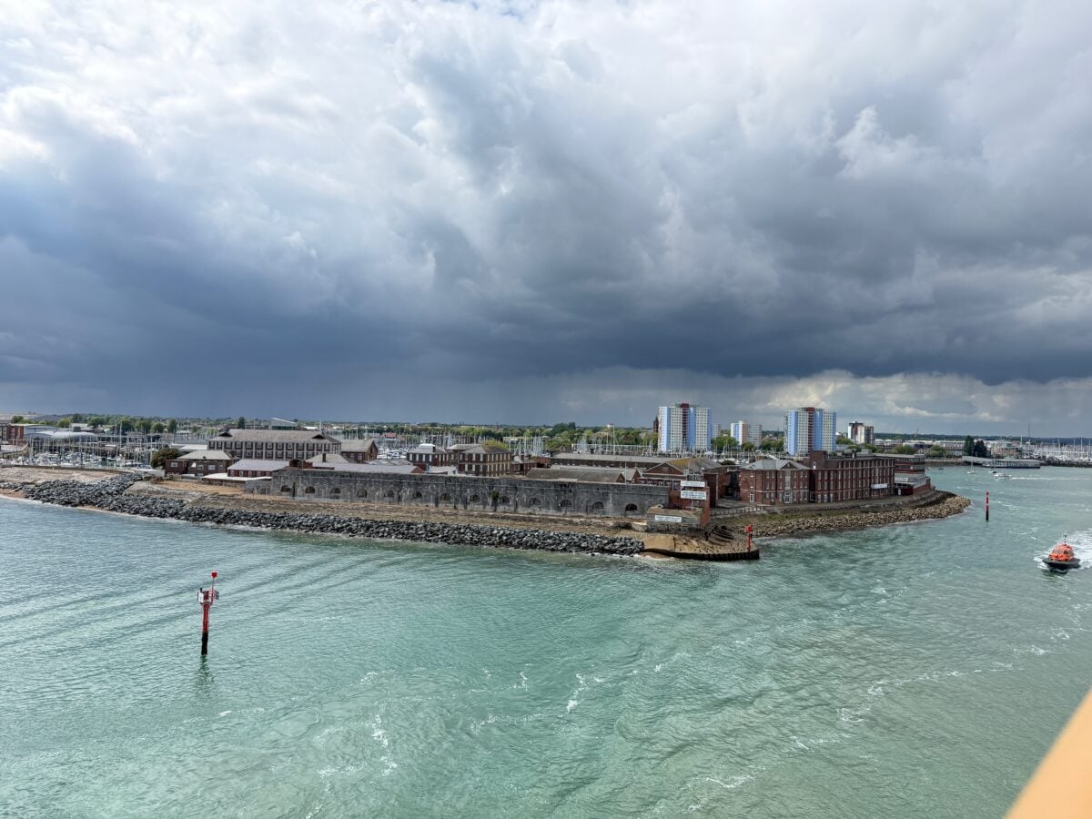 A coastal cityscape features old brick buildings on a narrow, rocky peninsula surrounded by greenish water under a dramatic cloudy sky, with a few red buoys and a small boat on the right.