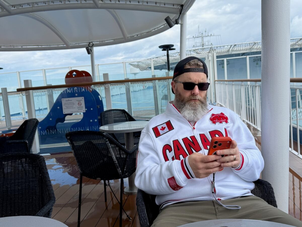 A bearded man wearing sunglasses, a backwards cap, and a white Canada jacket sits on a cruise ship deck using a red smartphone. Empty chairs and a blue waterslide are visible in the background.