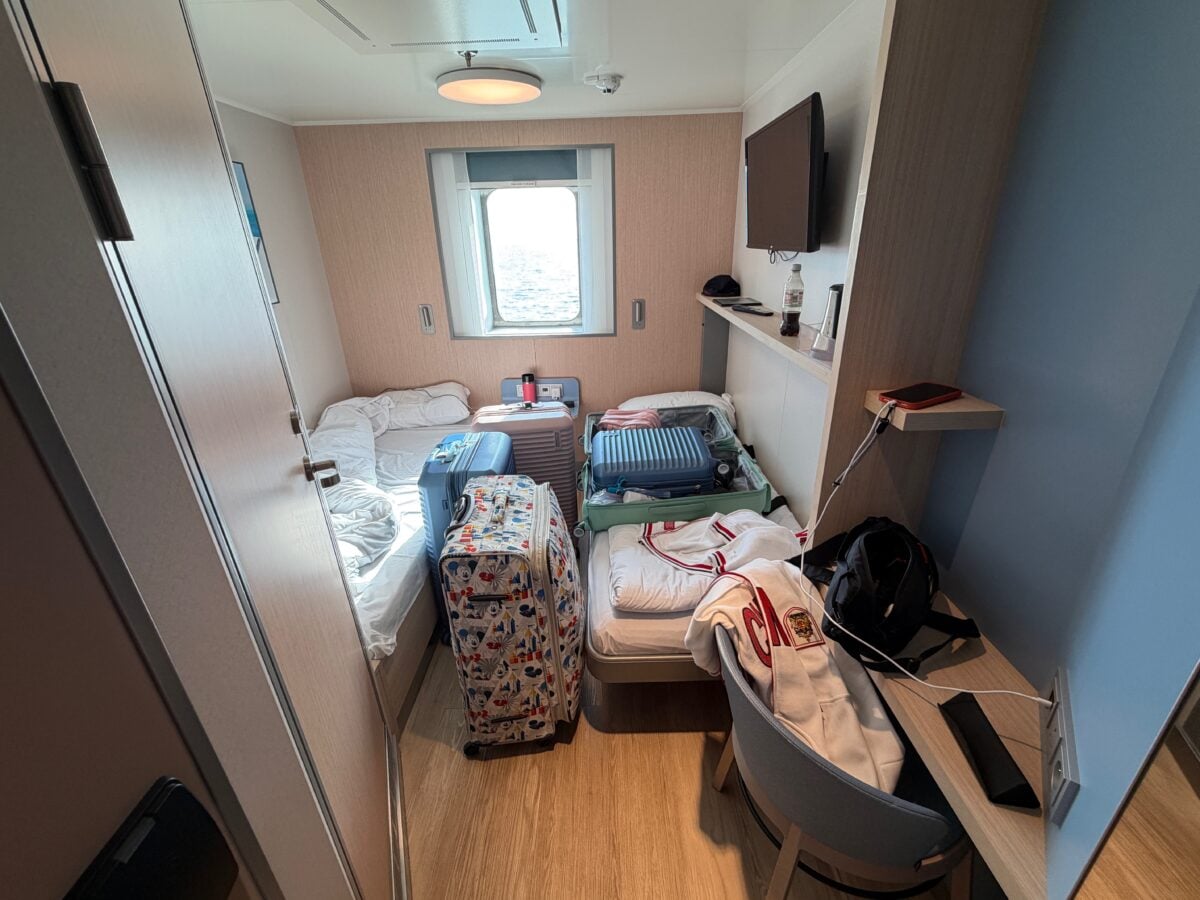 A small, tidy cruise ship cabin with two unmade single beds, a window with an ocean view, luggage, bags, and clothes on the beds, a TV on the wall, and a desk with electronics and other personal items.