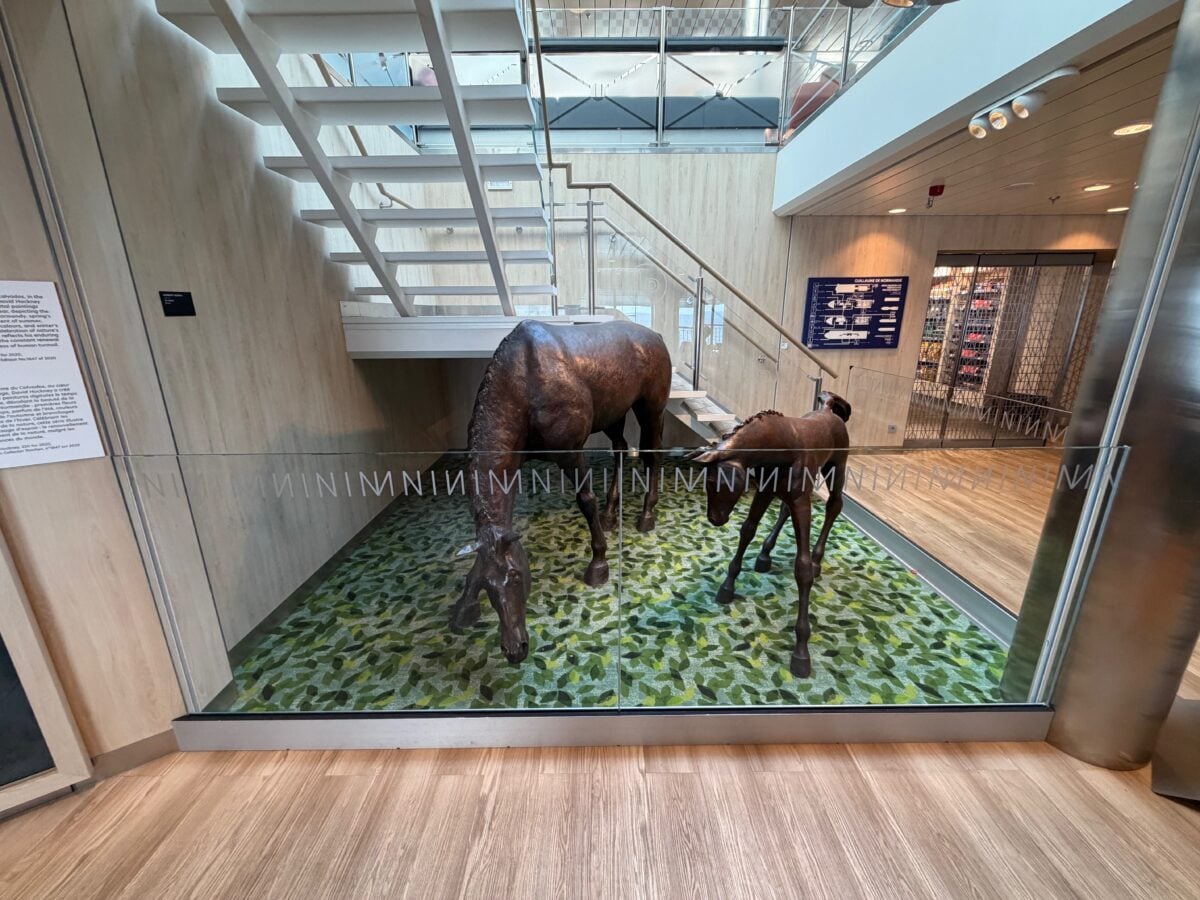 Two life-sized horse sculptures, an adult and a foal, stand on a green patterned floor under a modern staircase in a glass-walled display area inside a building.