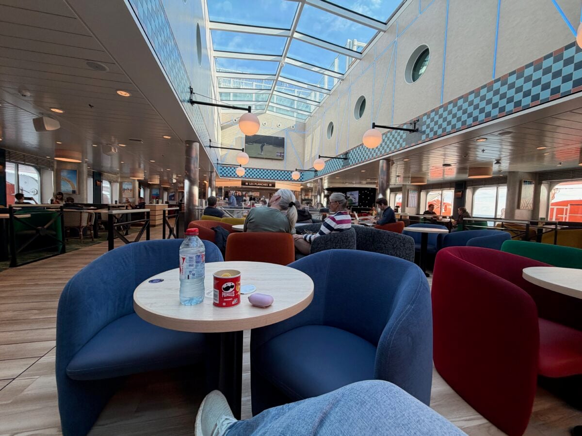 People sit and relax in a brightly lit ferry lounge with colorful chairs and tables. A bottle of water, a Pringles can, and a small object are on the table in the foreground. Large windows and a glass ceiling let in sunlight.