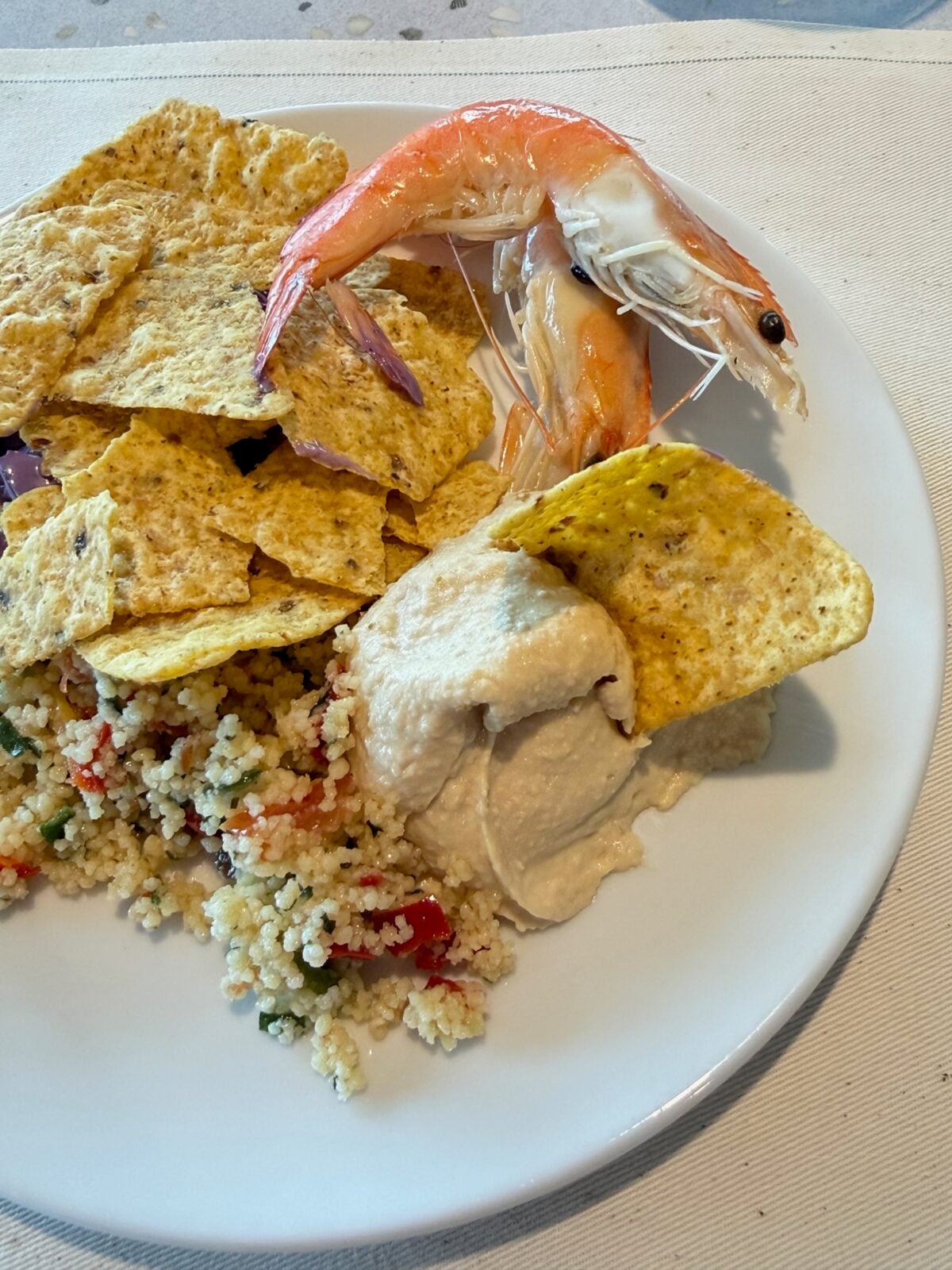 A white plate with tortilla chips, a scoop of hummus, a couscous salad with vegetables, and a whole cooked shrimp on top. The plate is set on a light-colored tablecloth.