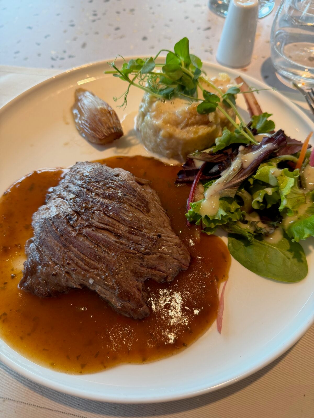 A plated meal with a piece of grilled steak in brown sauce, mashed potatoes with greens on top, a roasted shallot, and a small green salad with dressing, served on a white plate.