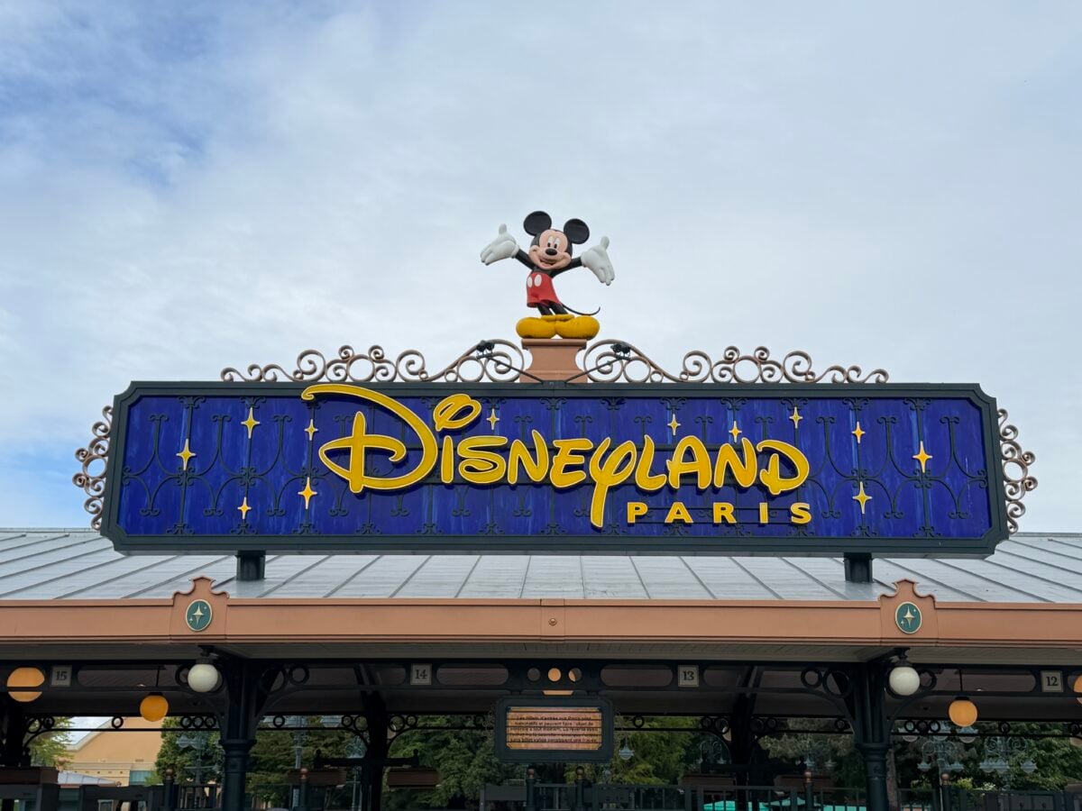 The entrance sign for Disneyland Paris features a large, cheerful Mickey Mouse statue standing on top. The sign has a blue background with yellow stars and bold yellow lettering.