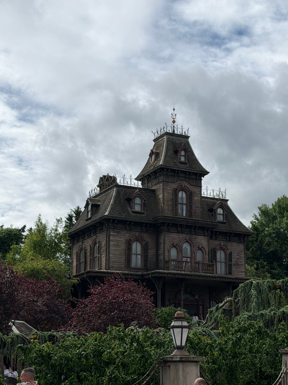 A large, dark, Victorian-style haunted house with ornate details stands behind lush greenery and trees under a cloudy sky.