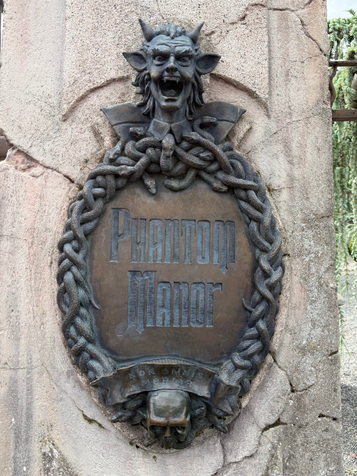 A bronze plaque on a stone wall reads Phantom Manor, bordered by an ornate, twisted frame. Above the plaque is a screaming demonic face, and below is a skull. The plaque has an eerie, gothic style.