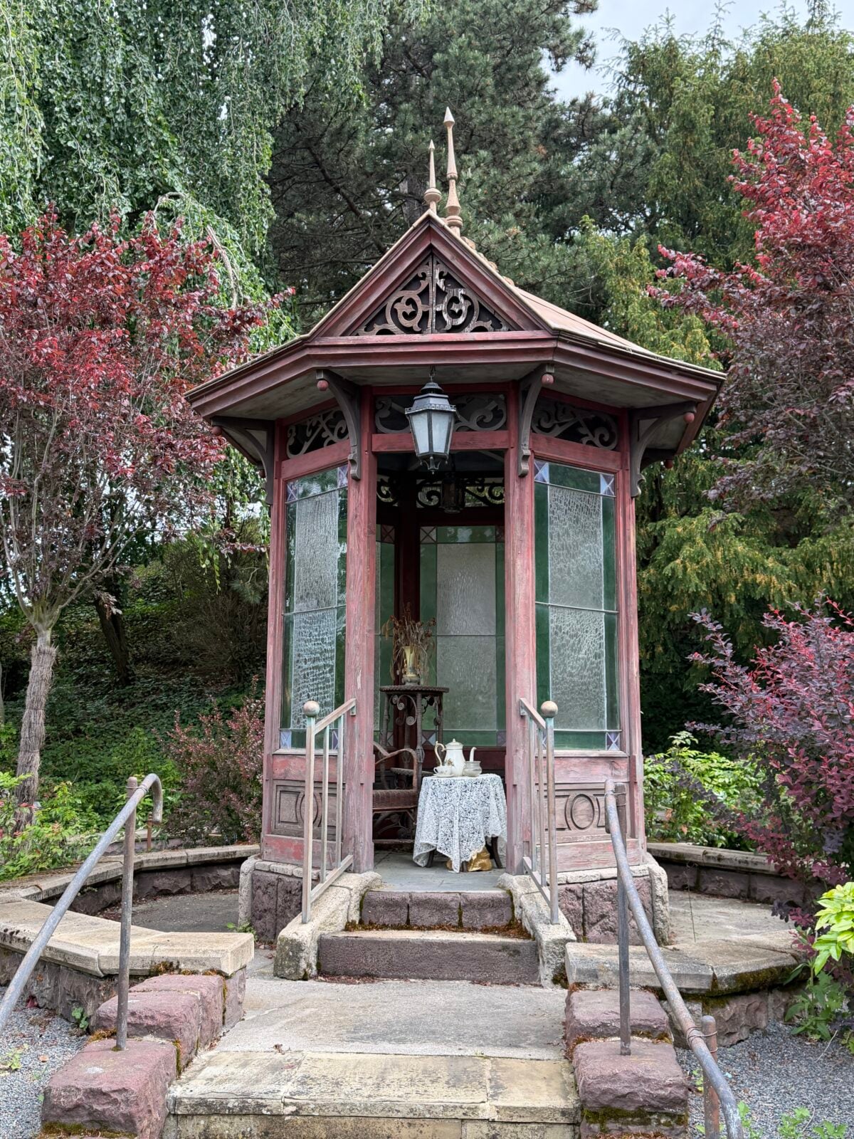 A small, ornate wooden gazebo with decorative trim and frosted glass panels stands among lush trees and shrubs. Inside, a table with a white lace cloth and chairs are visible. Stone steps and railing lead to the entrance.
