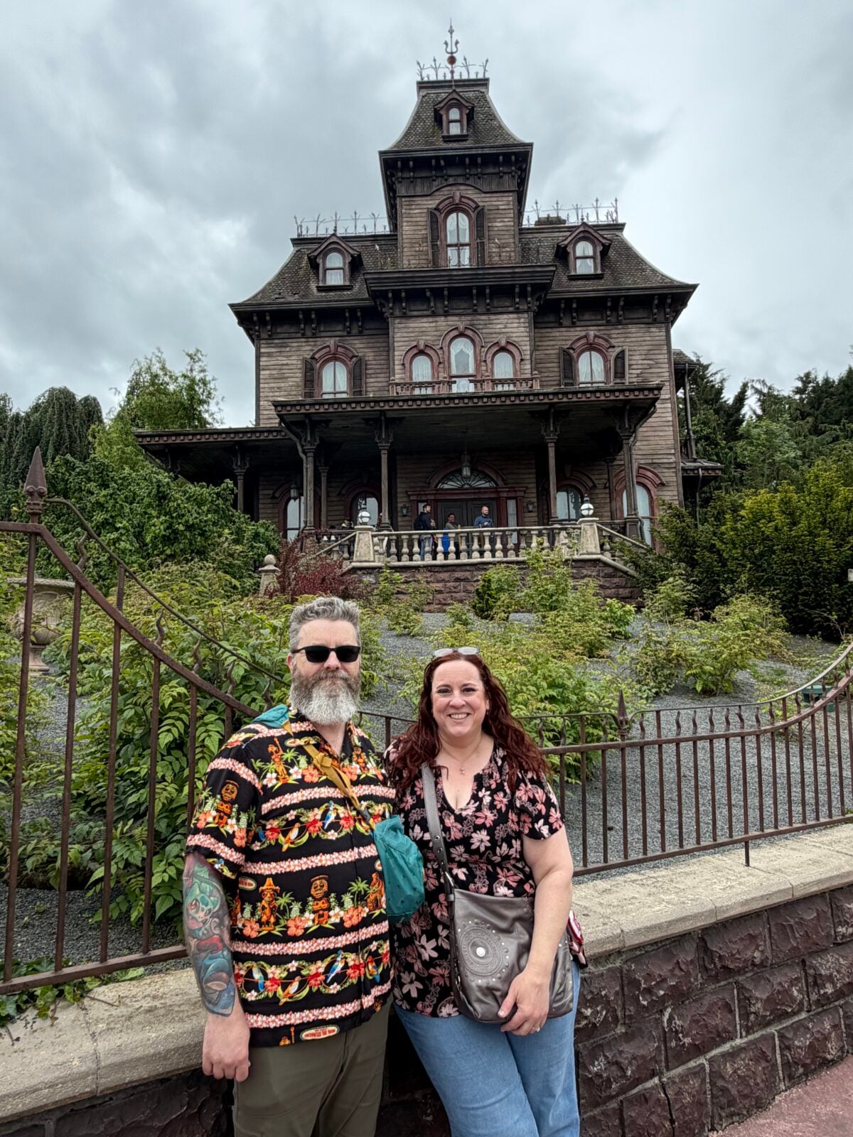 A man and woman stand smiling in front of a large, old, Victorian-style haunted house with iron railings and cloudy skies above.