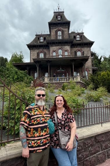 A man and woman stand smiling in front of a large, old, Victorian-style haunted house with iron railings and cloudy skies above.