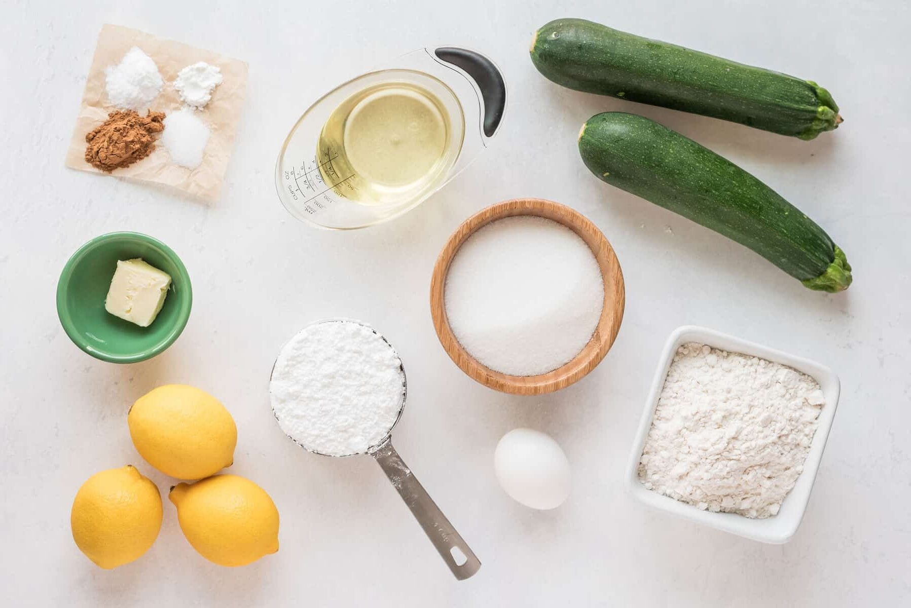Several baking ingredients are arranged on a white surface, including two zucchinis, three lemons, an egg, a bowl of sugar, a bowl of flour, powdered sugar, butter, oil, cinnamon, baking powder, and baking soda.