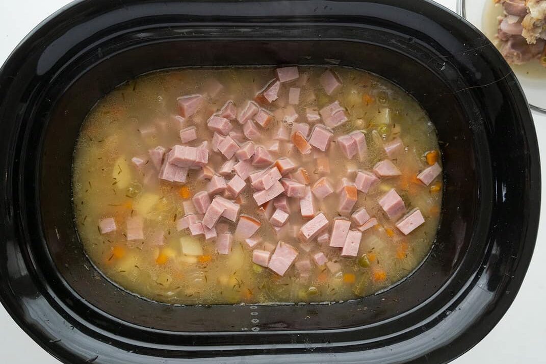 A slow cooker filled with soup containing diced ham, carrots, potatoes, and broth. The ingredients are visible and appear to be cooking together.