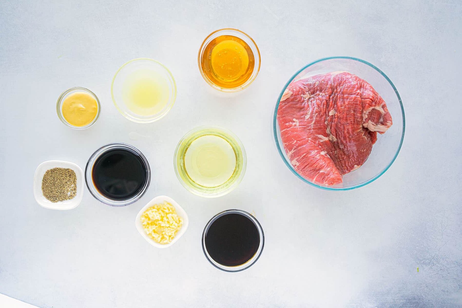 A glass bowl with raw beef and small bowls containing honey, soy sauce, vinegar, lemon juice, mustard, black pepper, minced garlic, and a dark sauce, arranged on a light gray surface.