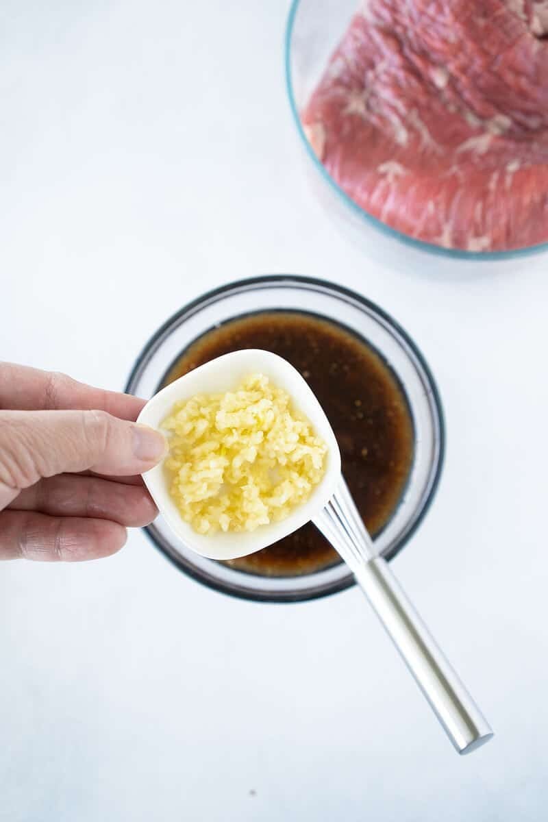 A hand holds a small white dish of minced garlic above a bowl of dark marinade with a whisk in it; a bowl of raw sliced beef is partially visible in the background.