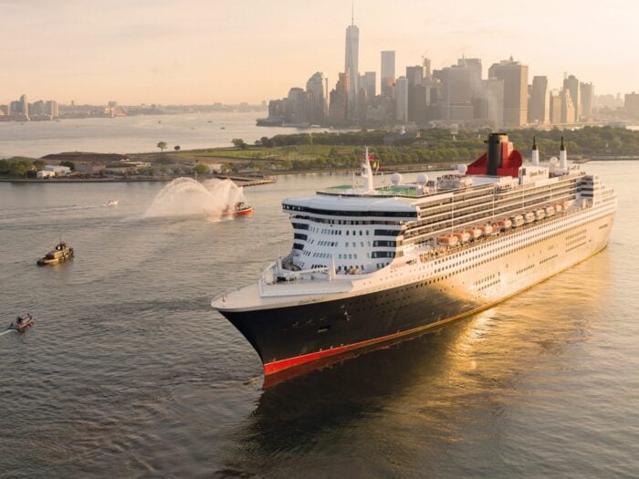 A large cruise ship sails on a river toward a city skyline at sunset, with water spray from a tugboat and skyscrapers visible in the background.
