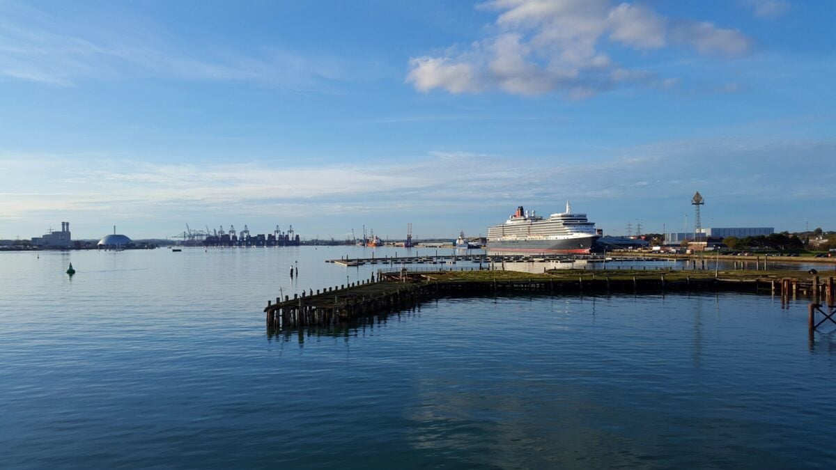 A large cruise ship is docked at a harbor with calm blue water, industrial cranes, and buildings visible in the background under a partly cloudy sky.