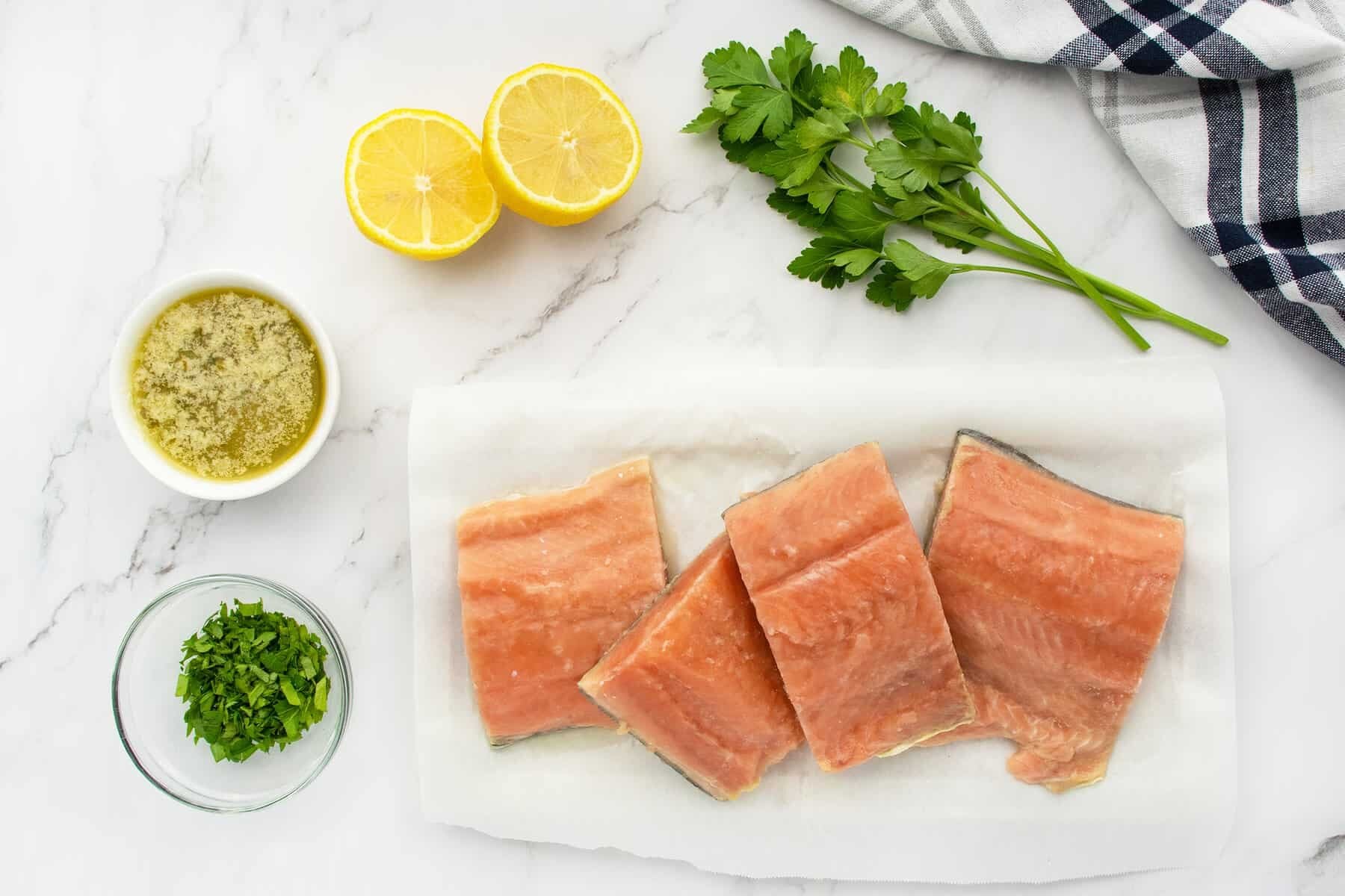 Four raw salmon fillets on parchment paper, fresh parsley, two lemon halves, melted butter with herbs, chopped parsley in a bowl, and a checkered towel on a marble surface.