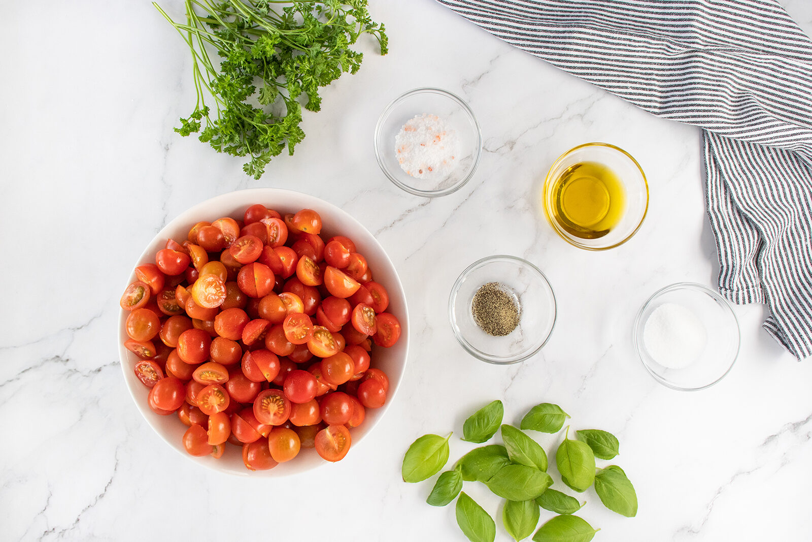A bowl of halved cherry tomatoes, fresh basil leaves, parsley, bowls of olive oil, salt, and pepper are arranged on a white marble surface with a striped cloth on the side.