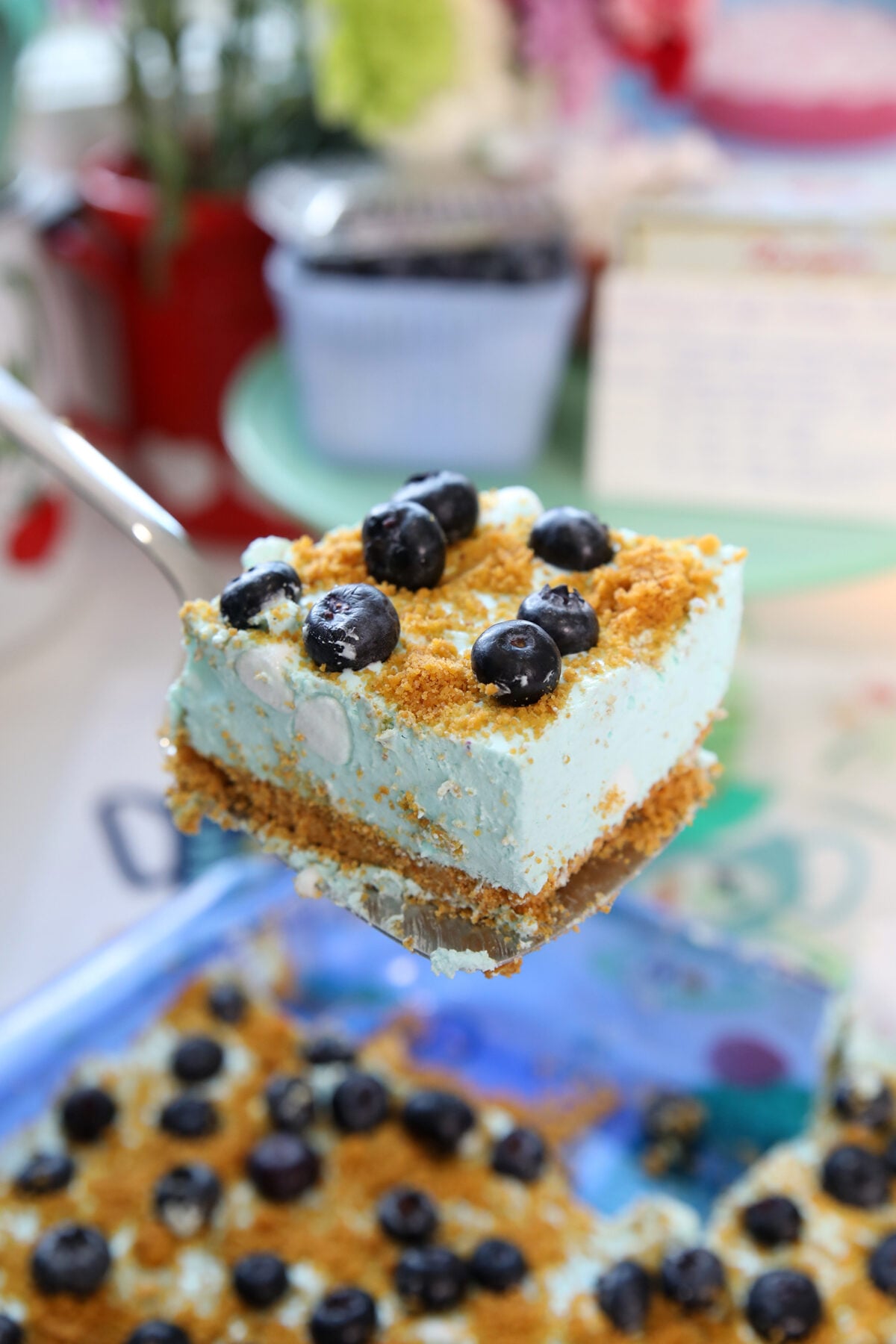 A spatula lifts a square of creamy, blue dessert topped with blueberries and crumbs. More pieces of the dessert and scattered blueberries are visible in the background.