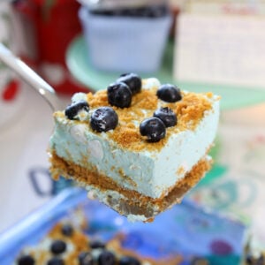 A spatula lifts a square of creamy, blue dessert topped with blueberries and crumbs. More pieces of the dessert and scattered blueberries are visible in the background.