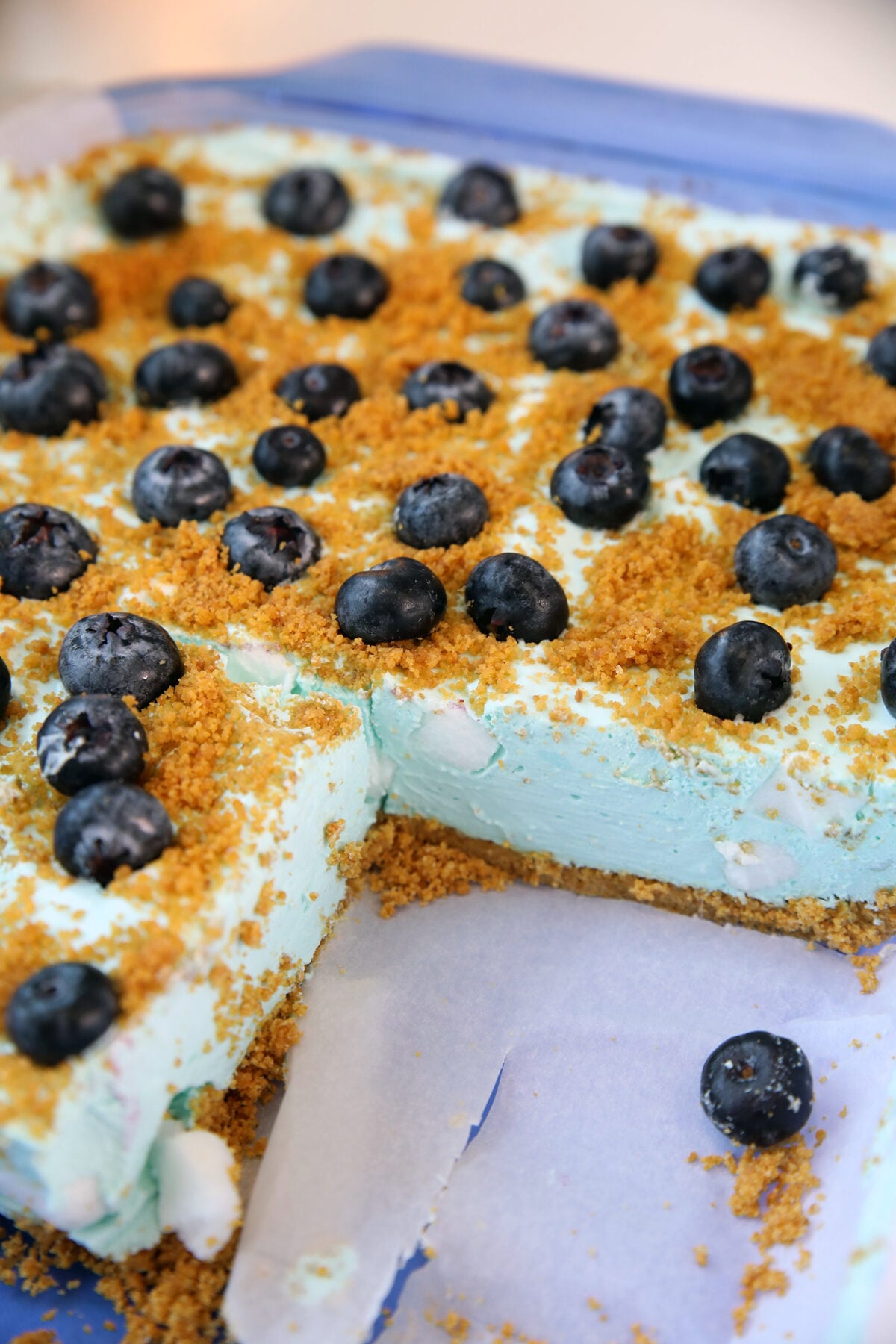 A square dessert with a creamy, light blue filling, topped with fresh blueberries and crumbled golden crust, sits on parchment paper with one piece removed.