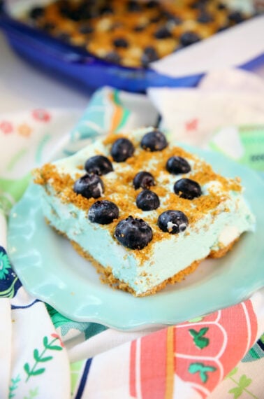 A slice of creamy blueberry dessert with a graham cracker crust, topped with fresh blueberries, sits on a light blue plate over a colorful patterned cloth. The rest of the dessert is visible in a blue dish in the background.