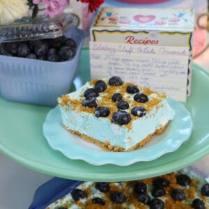 A slice of blueberry no-bake cheesecake topped with blueberries sits on a blue plate. Behind it are fresh blueberries, a handwritten recipe card, flowers, and a lit candle.