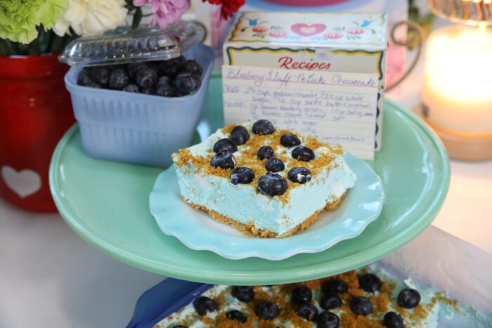 A slice of blueberry no-bake cheesecake topped with blueberries sits on a blue plate. Behind it are fresh blueberries, a handwritten recipe card, flowers, and a lit candle.