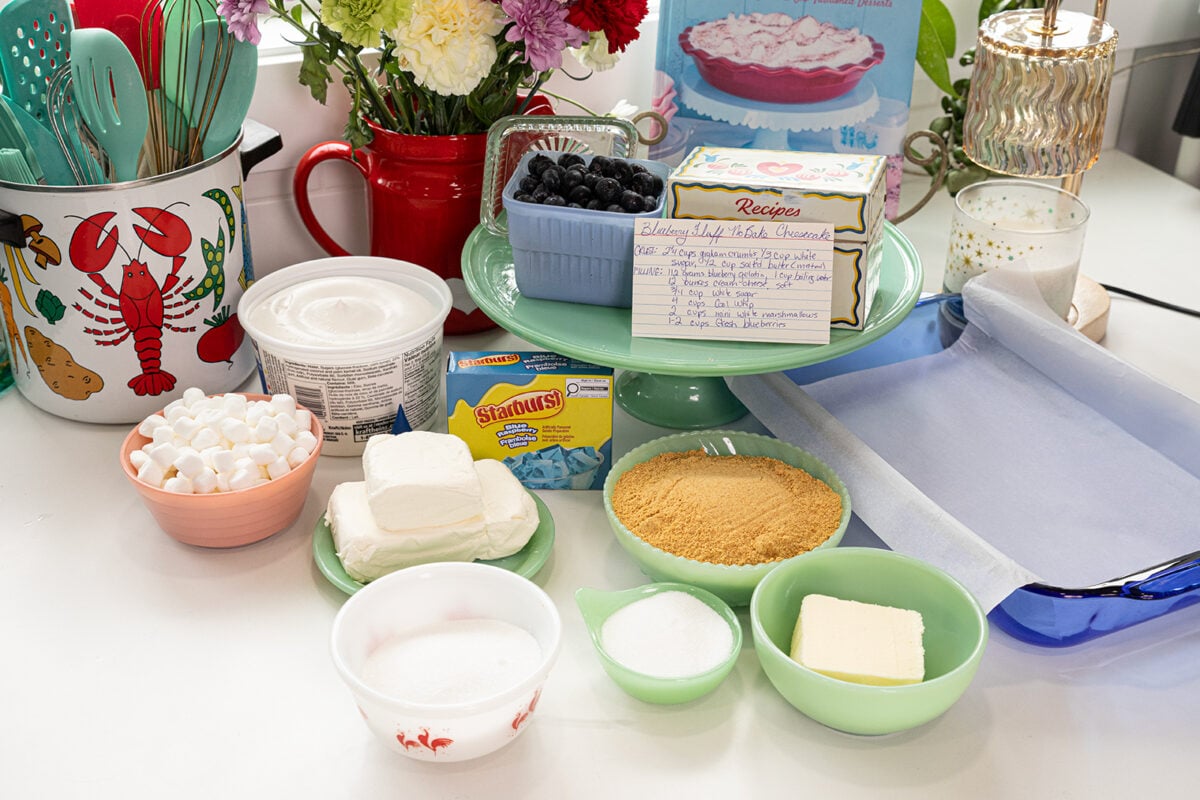 Baking ingredients on a counter include cream cheese, marshmallows, sugar, graham cracker crumbs, butter, whipped cream, blueberries, a recipe card, and kitchen utensils. Flowers and decor are in the background.