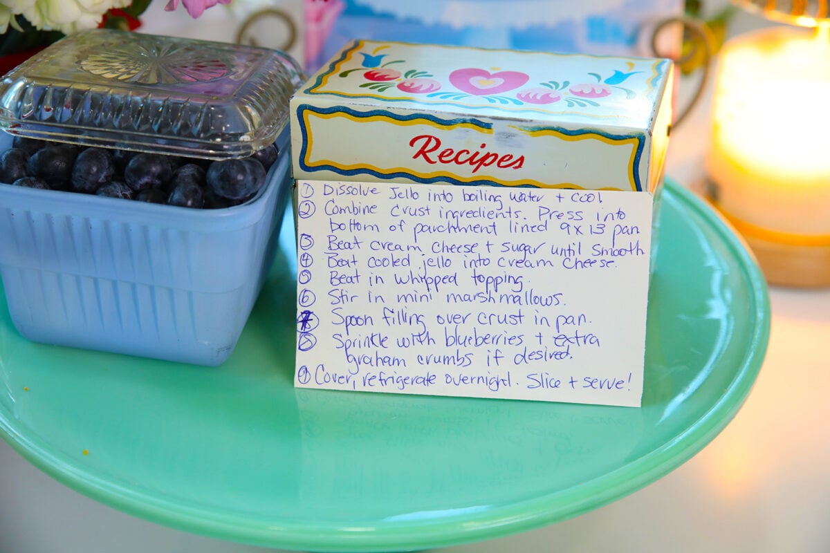 A handwritten recipe card next to a container of blueberries and a decorative recipe box, all placed on a light blue cake stand with a candle in the background.