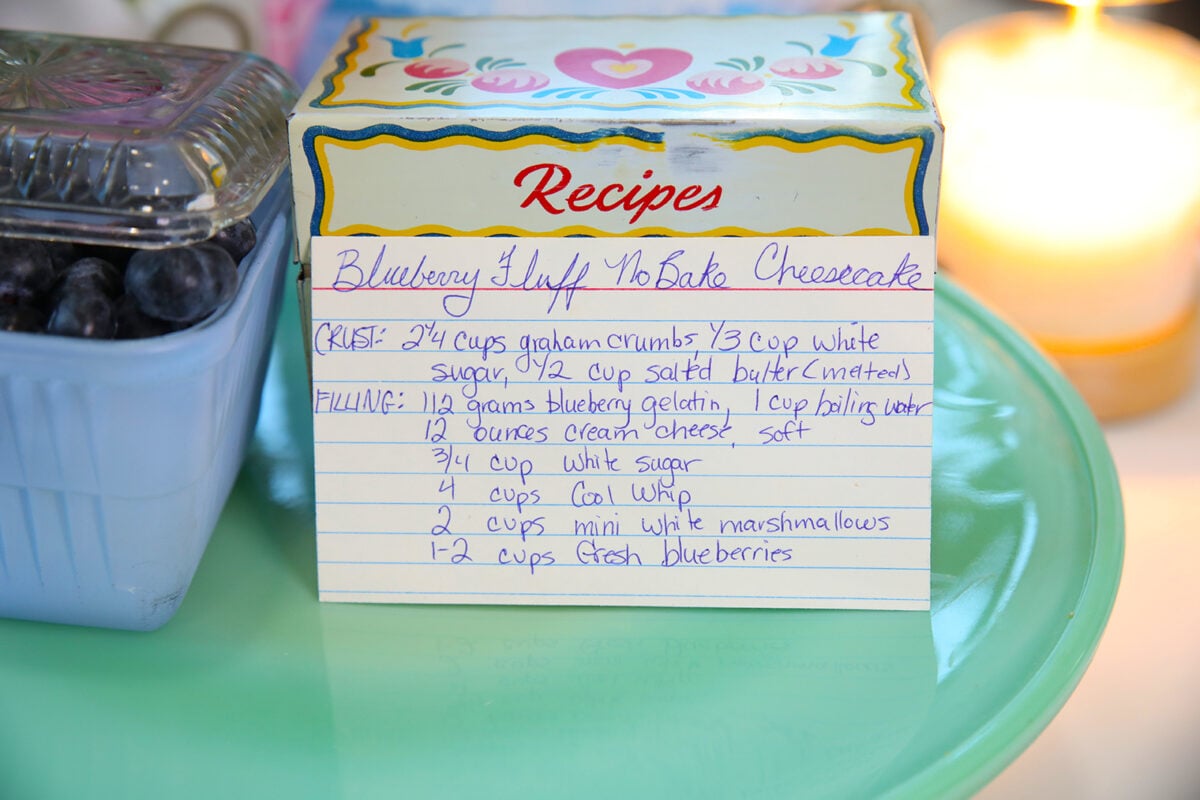A handwritten recipe card for Blueberry Fluff No-Bake Cheesecake sits on a blue plate with a blueberries container, a decorative recipe box, and a lit candle nearby.