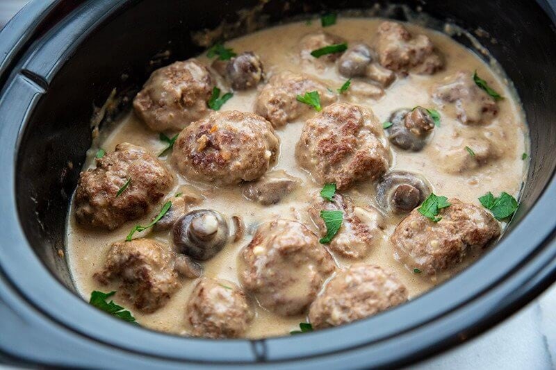 Meatballs in creamy sauce with mushrooms and parsley, cooked in a black slow cooker.