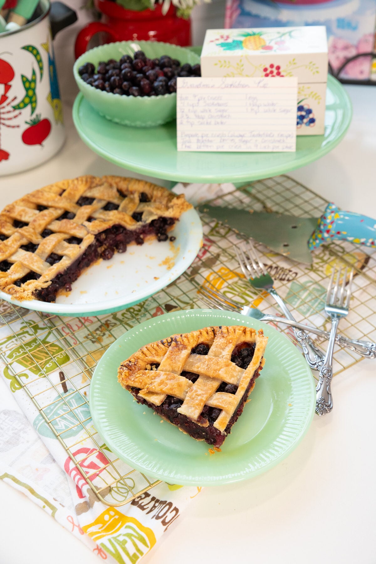 A slice of lattice-topped blueberry pie on a green plate sits in front of a partially eaten pie, with forks, a bowl of blueberries, and a handwritten recipe card on a colorful kitchen counter.