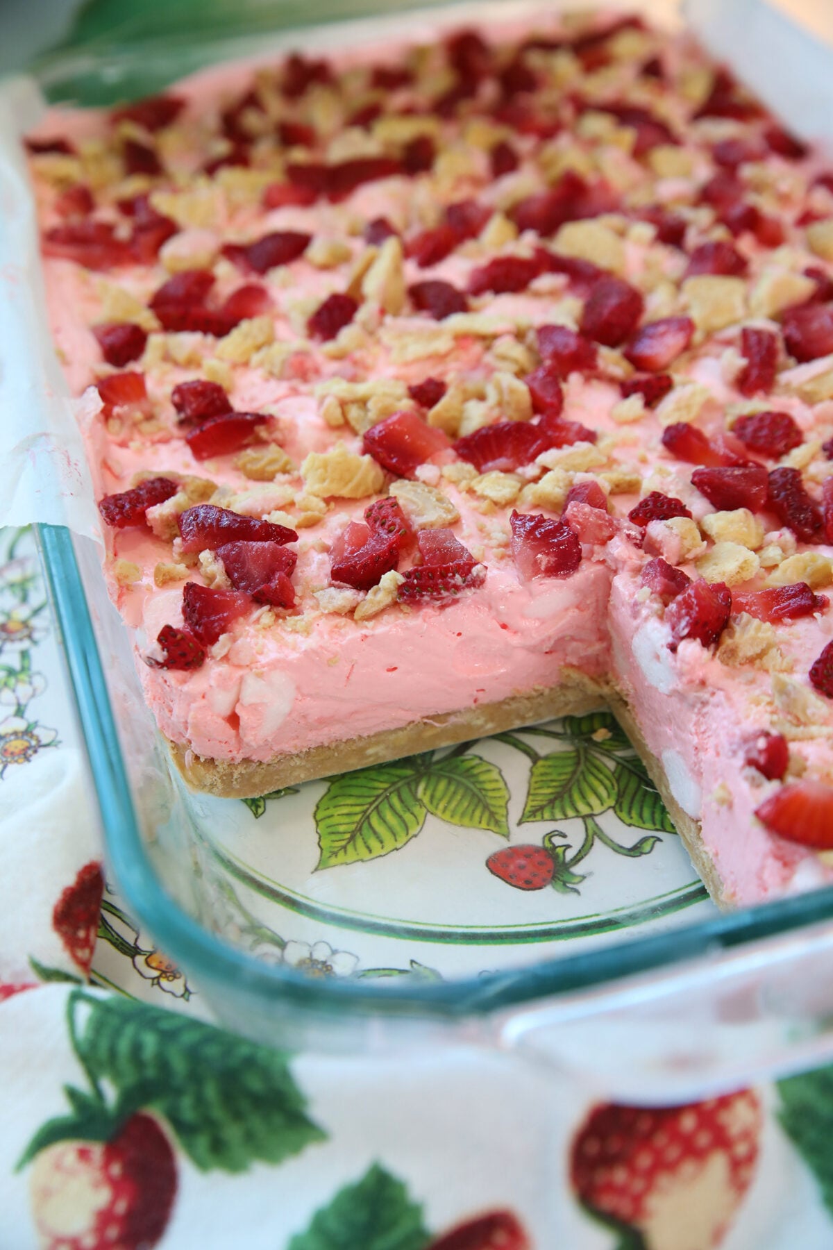 A glass dish filled with a pink dessert topped with chopped strawberries and nuts, with one large rectangular piece missing. The dish sits on a strawberry-patterned cloth.