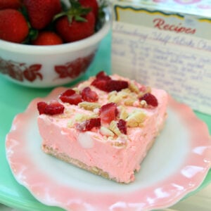 A slice of pink dessert topped with strawberries and nuts on a pink plate, with a bowl of strawberries and a handwritten recipe card in the background.