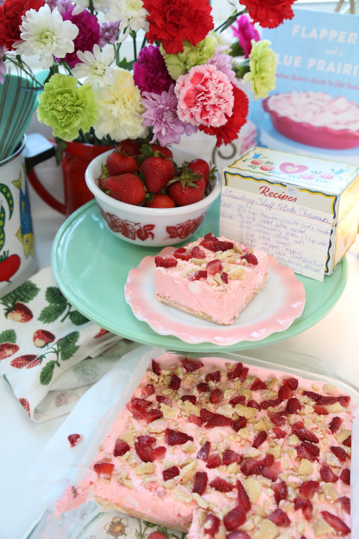A pink dessert with strawberries sits on a plate, with a bowl of strawberries, a floral towel, handwritten recipe cards, and a bouquet of colorful flowers in the background.