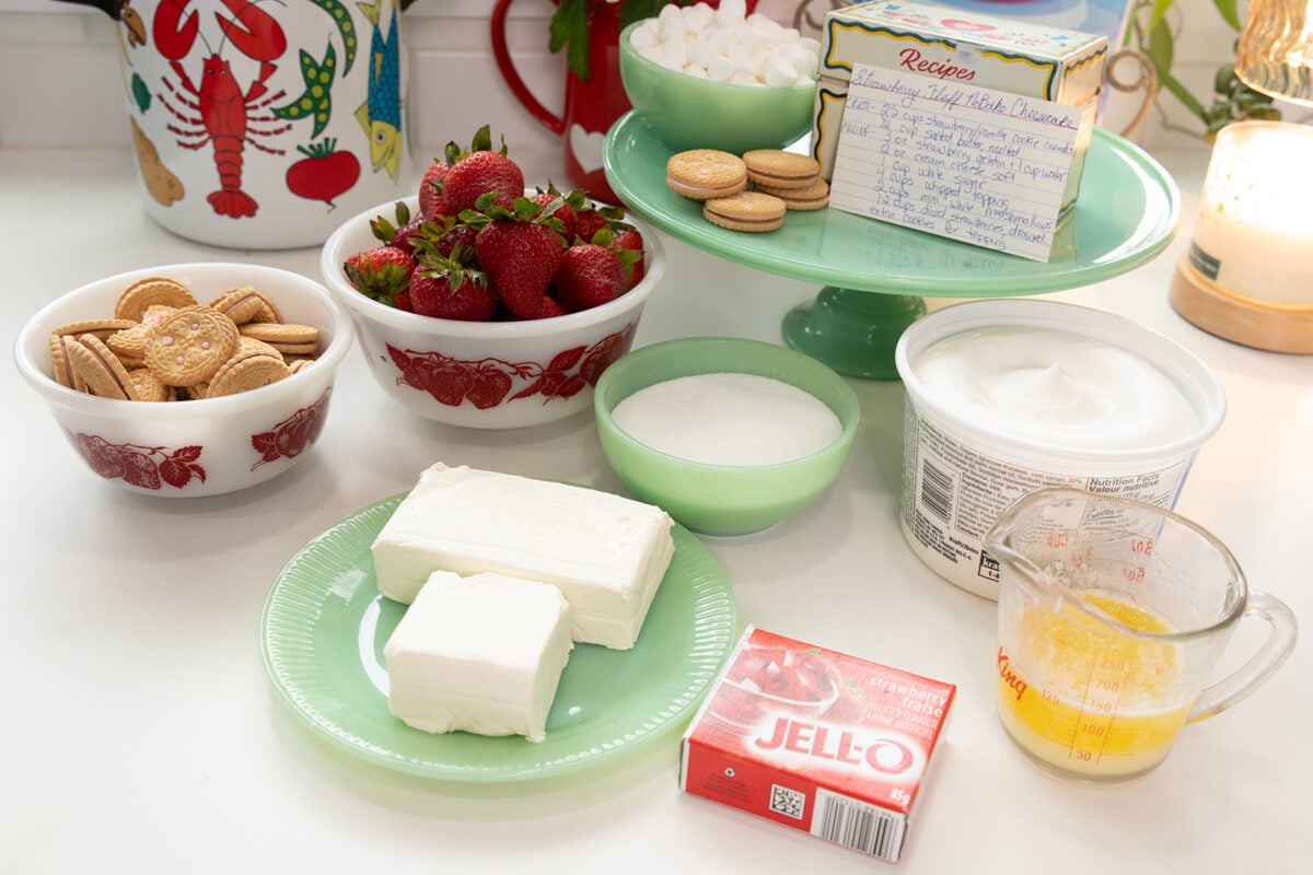 A countertop with bowls of strawberries, golden sandwich cookies, sugar, cream cheese, Cool Whip, melted butter, a box of strawberry Jello, and a recipe box, arranged for making a dessert.