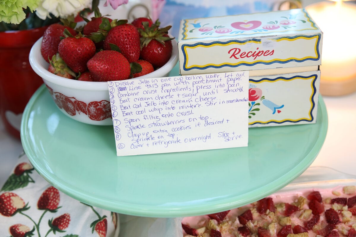 A handwritten recipe card sits on a teal cake stand beside a bowl of fresh strawberries and a vintage recipe box decorated with hearts and flowers. Strawberry dessert bars are partially visible in the foreground.