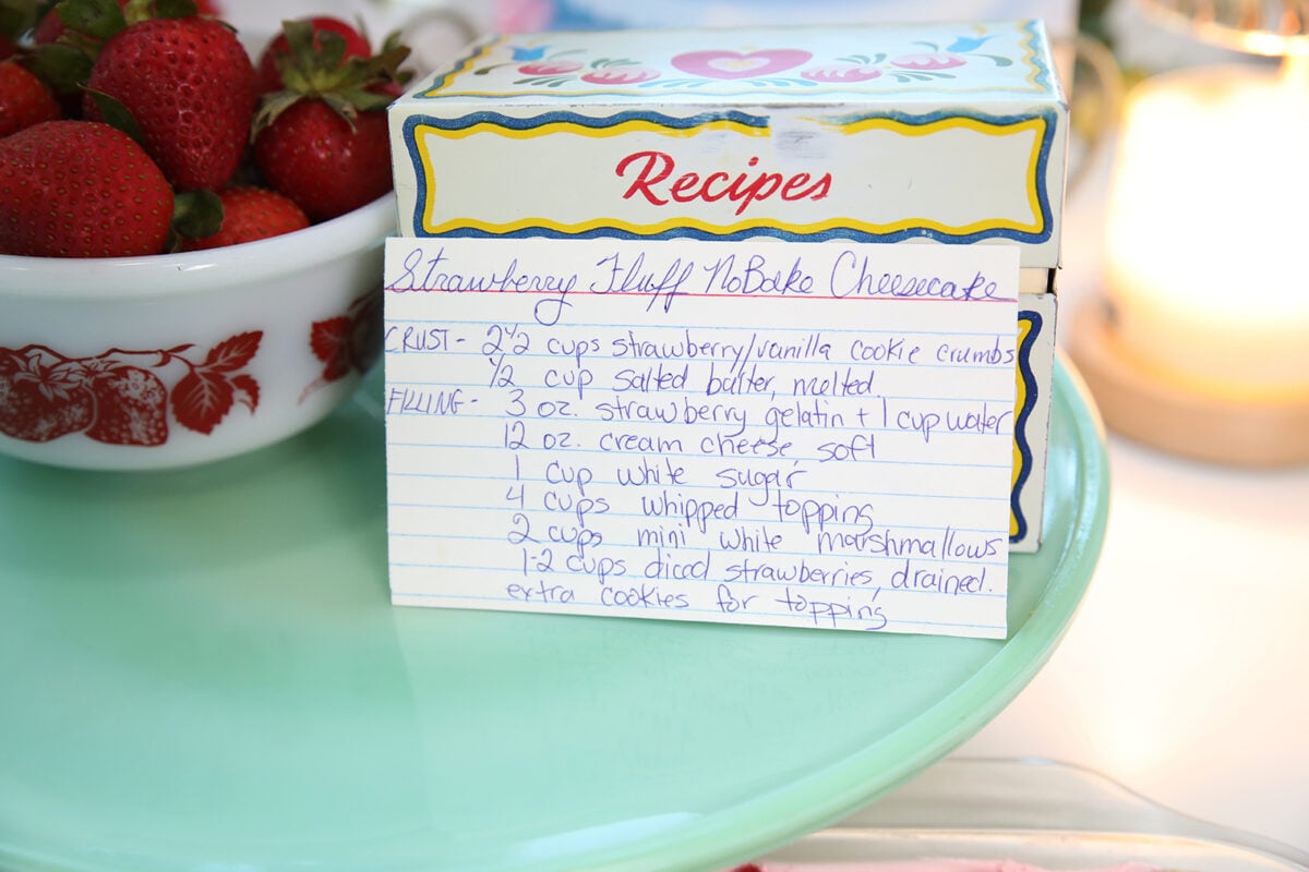 A handwritten recipe card for Strawberry Fluff No-Bake Cheesecake sits on a blue tray, next to a bowl of fresh strawberries. The recipe box is colorful and decorated with hearts and flowers.