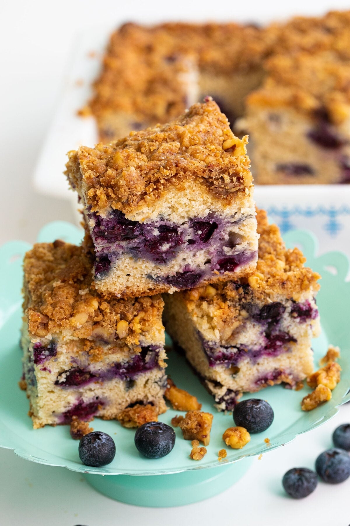 Three squares of blueberry coffee cake with crumb topping are stacked on a decorative plate, with fresh blueberries scattered around and more cake visible in the background.