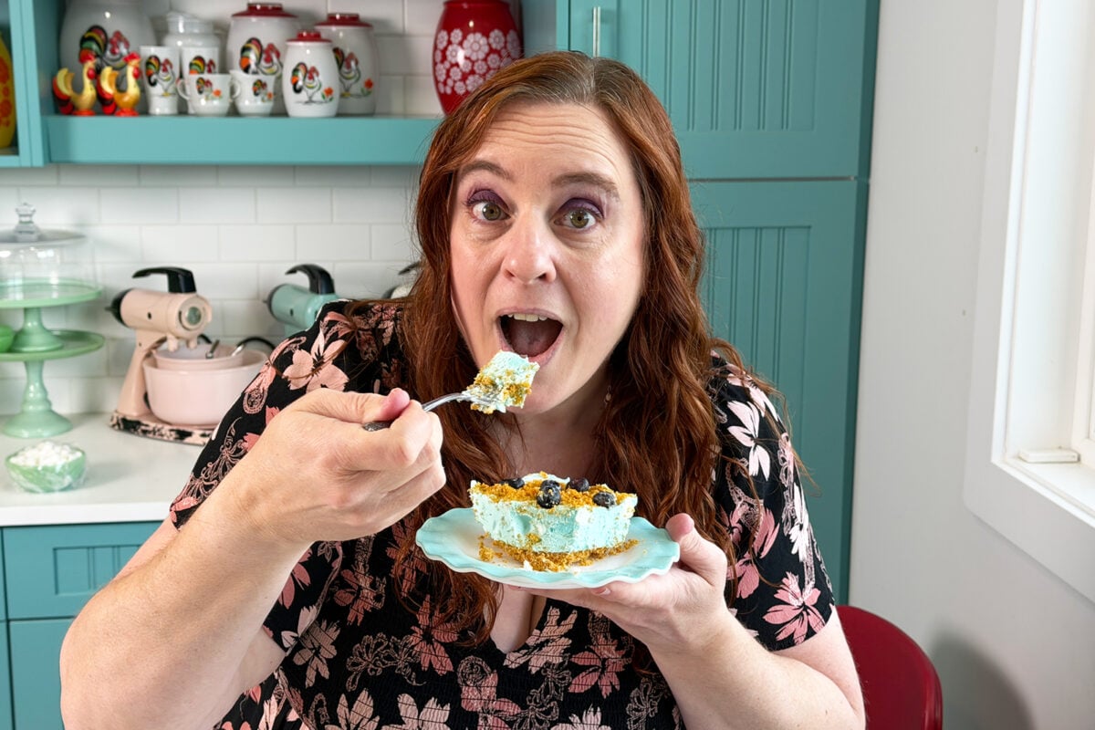 A woman with long brown hair and a floral top holds a plate with blue dessert, about to take a bite with a fork, sitting in a colorful kitchen with teal cabinets and decorative dishes.