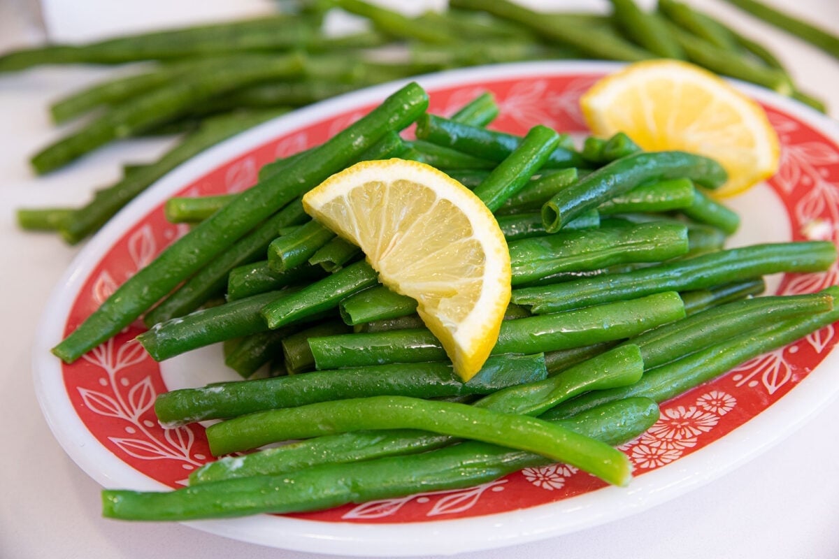 A red and white plate filled with steamed green beans, garnished with a lemon wedge. More green beans are visible in the background.