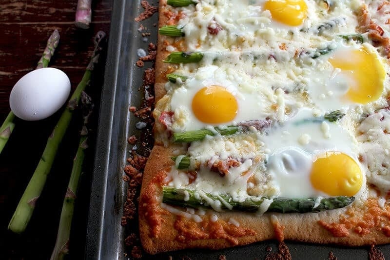 Rectangular pizza topped with melted cheese, asparagus spears, and sunny-side-up eggs on a baking tray. An egg and asparagus spears rest on the dark wooden table beside the tray.