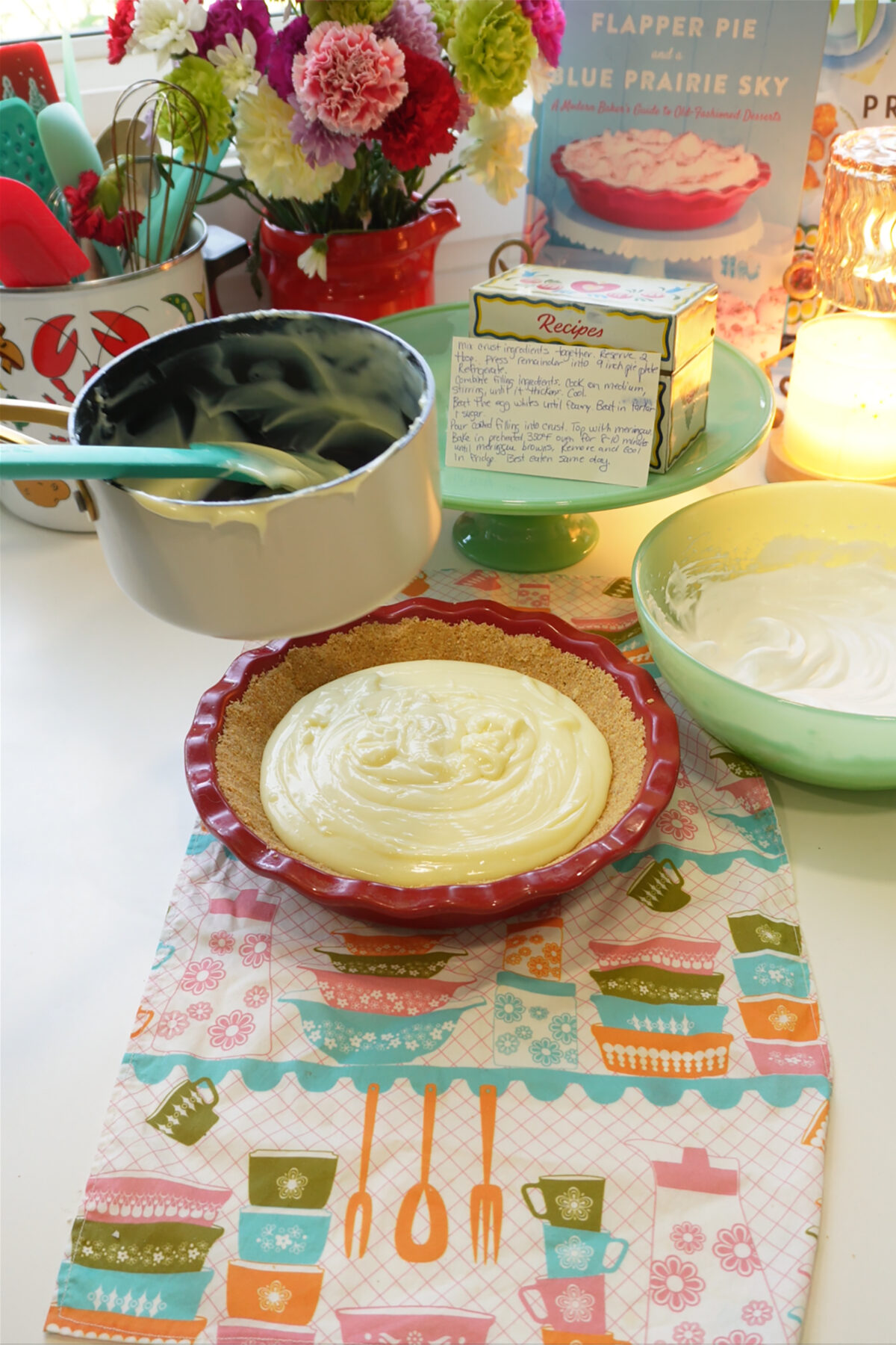 A pie dish with creamy filling sits on a colorful cloth. Nearby are a bowl with a spatula, a whipped mixture in a green bowl, flowers, a candle, and a recipe card. A cookbook stands in the background.