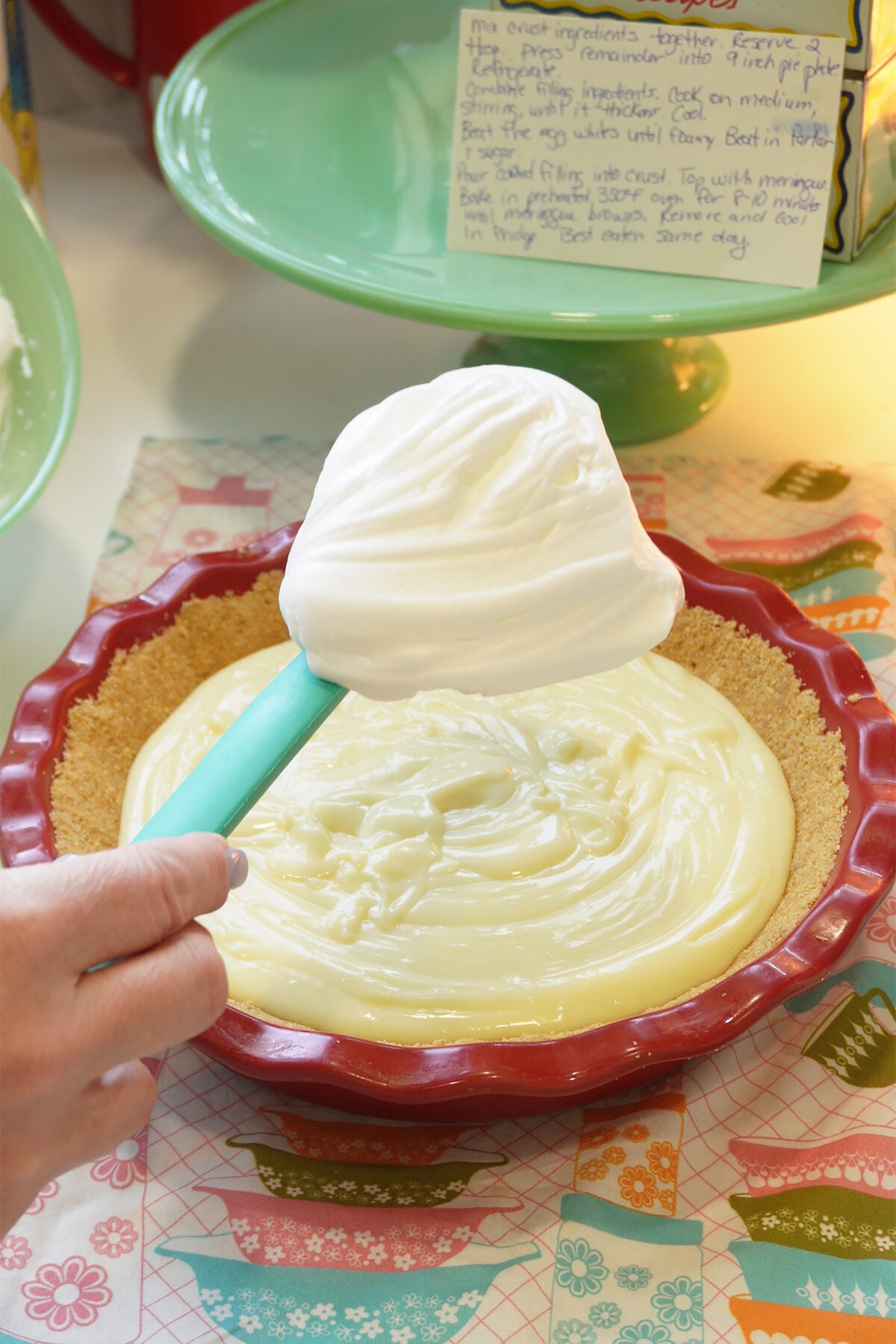 A hand spreads fluffy whipped topping over a creamy pie filling in a graham cracker crust, with a handwritten recipe card and colorful printed napkin nearby.
